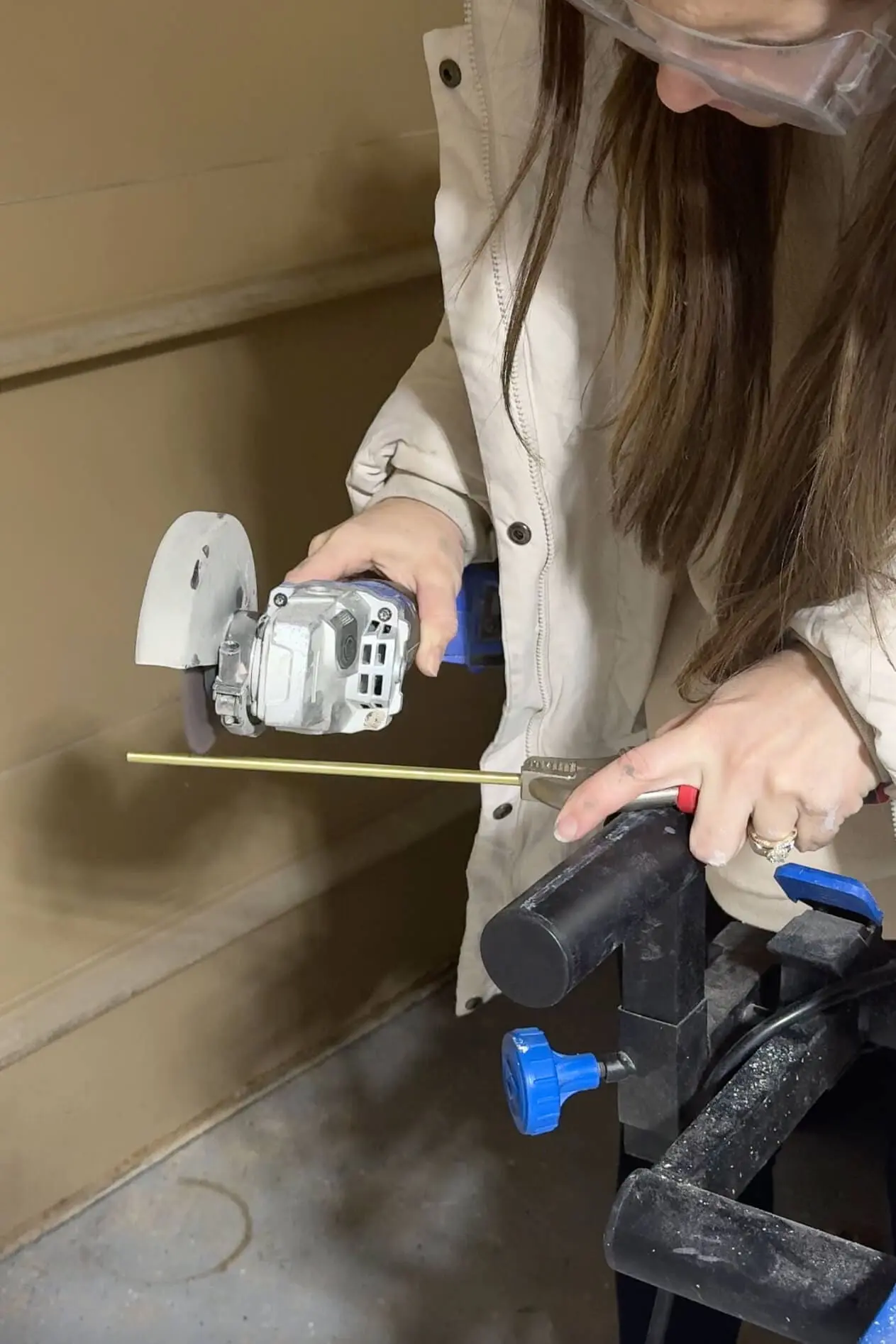 A close-up of a woman using a power tool to cut a thin brass rod, held steady with clamps on a workbench in a garage setting.